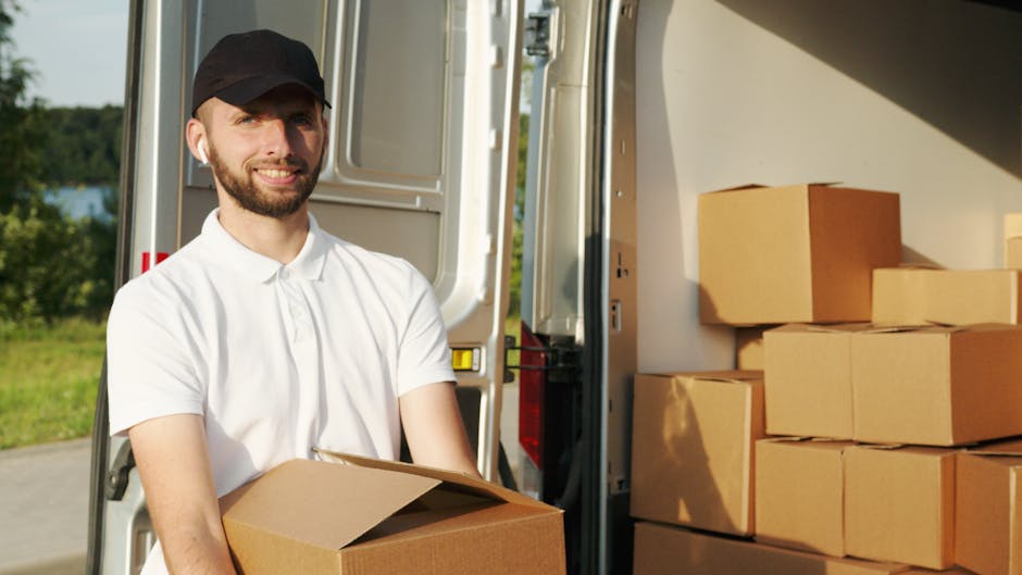 A man dressed in a red and navy jacket, black trousers, and a blue beanie hat is sitting on the open tailgate of a black van with a spacious cargo area, which contains several large brown cardboard bo