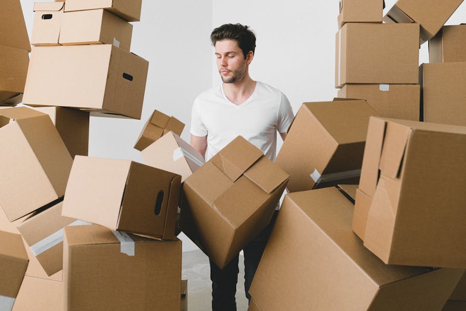 A man with dark hair and a light complexion, dressed in a plain white t-shirt, is standing amidst numerous brown cardboard boxes of varying sizes, some with visible white packing tape, inside a well-lit indoor space with white walls. He appears to be inspecting or arranging the boxes, which are stacked around him and some are suspended in mid-air, suggesting they are being lifted or moved. The boxes are likely used for packing household items during a home relocation or for storage purposes. Nearby, a doorway leads to another room, and a portion of a moving van can be seen outside through the open door, indicating the process of loading or unloading cargo. The environment reflects the packing and moving process, typical of professional removal services such as those offered by Man with Van Harold Park, who specialise in house removals, furniture transport, and last-minute storage options, especially in cases of last-minute cancellations or delays in the relocation timeline.