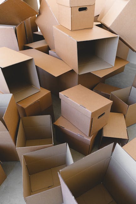 A collection of open and closed cardboard boxes of various sizes, some with handles, are piled inside a home, likely part of a packing process during a house move. The boxes are arranged on a floor with some stacked on top of each other and others scattered around, indicating recent packing or preparation for transportation. The environment appears to be an indoor space, possibly a hallway or storage area, with neutral lighting. This scene captures the logistics involved in furniture transport and packing as part of home relocation services offered by Man with Van Harold Park, emphasizing the use of cardboard containers for efficiently organizing personal belongings during packing and loading stages of a move.
