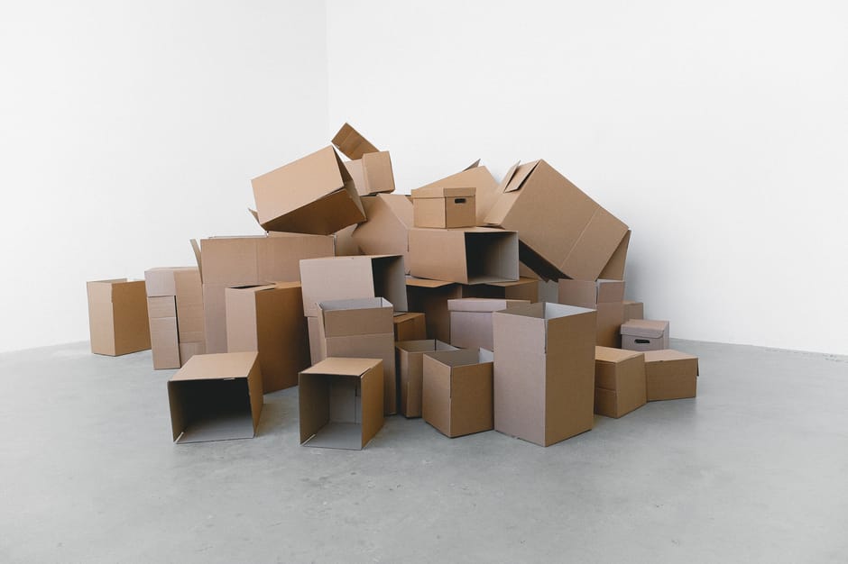 A man with dark hair and a light complexion, dressed in a plain white t-shirt, is standing amidst numerous brown cardboard boxes of varying sizes, some with visible white packing tape, inside a well-lit indoor space with white walls. He appears to be inspecting or arranging the boxes, which are stacked around him and some are suspended in mid-air, suggesting they are being lifted or moved. The boxes are likely used for packing household items during a home relocation or for storage purposes. Nearby, a doorway leads to another room, and a portion of a moving van can be seen outside through the open door, indicating the process of loading or unloading cargo. The environment reflects the packing and moving process, typical of professional removal services such as those offered by Man with Van Harold Park, who specialise in house removals, furniture transport, and last-minute storage options, especially in cases of last-minute cancellations or delays in the relocation timeline.