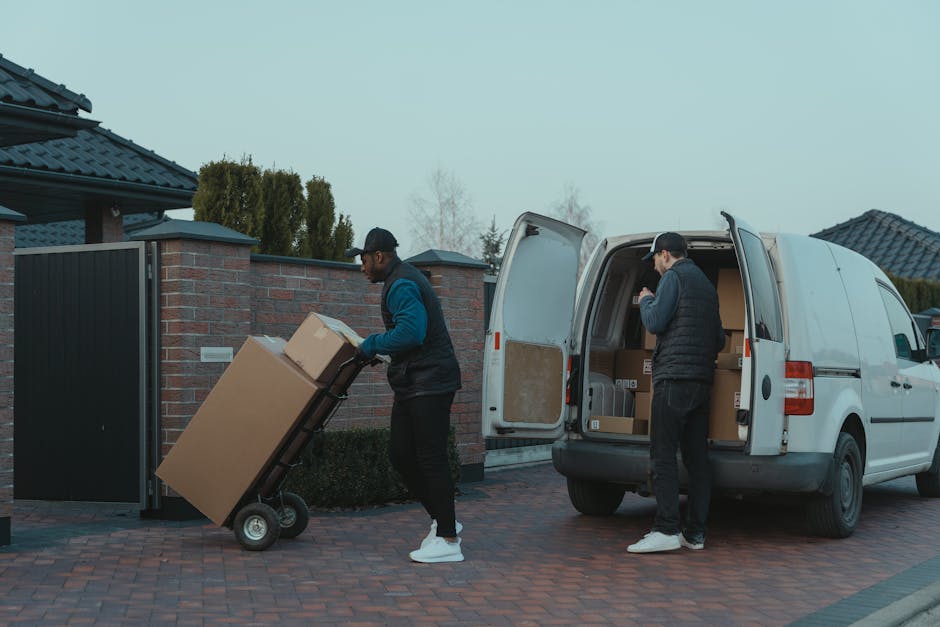 Two men are engaged in a home relocation process involving furniture and boxes loaded onto a white delivery van parked outside a residential building with brick walls and a tiled roof. One man, standi