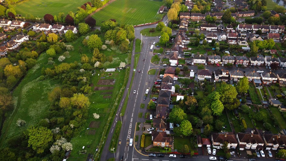An aerial view of a residential neighbourhood in Harold Park showing a two-lane road with several cars traveling along it, bordered by a well-maintained verge and sidewalks. To the right of the road, there is a dense array of terraced and semi-detached houses with front gardens, driveways, and parked vehicles, some with visible detached garages. To the left of the road, there is a large green park with numerous mature trees, some with white blossoms, open grassy areas, and a small section of vegetable plots or garden beds. The scene is captured in natural daylight with clear weather conditions, illustrating suburban living conditions. This setting is typical of a home relocation or furniture transport environment, where [COMPANY_NAME] might assist with various removals and moving services, including packing, loading, and transportation in the Harold Park area.