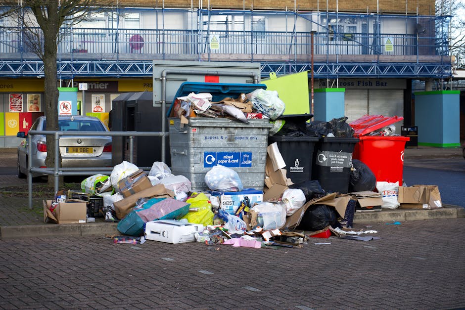 A collection of overflowing waste and recycling bins positioned on a paved sidewalk in front of a commercial building, with some cardboard boxes and plastic bags of rubbish spilling onto the ground. The bins include large gray mixed paper and cardboard containers, black recycling bins, and a red waste container, with additional black plastic bags piled nearby. The scene is outdoors, with a background featuring storefronts, a parking area with a silver car, and a building under construction or renovation covered in scaffolding and blue protective sheeting. Adjacent to the waste area is a metal barrier, and the overall setting suggests a typical urban environment where waste collection or disposal occurs, relevant to managing household or business rubbish during home relocation or moving processes, as handled by Man with Van Harold Park.