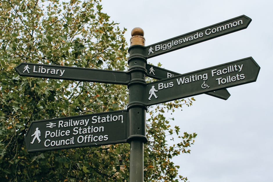A black multi-directional street signpost with white text and symbols, situated outdoors against a background of green foliage and an overcast sky. The signs point towards local landmarks and facilities including Biggleswade Common, the library, the bus waiting area, toilets, railway station, police station, and council offices. Each sign is attached to the post with metal brackets, and some include icons such as a walking figure, wheelchair accessibility, and a train symbol. The image captures a typical urban or village setting, useful for illustrating navigation and orientation or local amenities that might be relevant in the context of house removals or relocation services provided by Man with Van Harold Park, demonstrating the environment in which clients may seek assistance with home relocation or furniture transport within the local area.