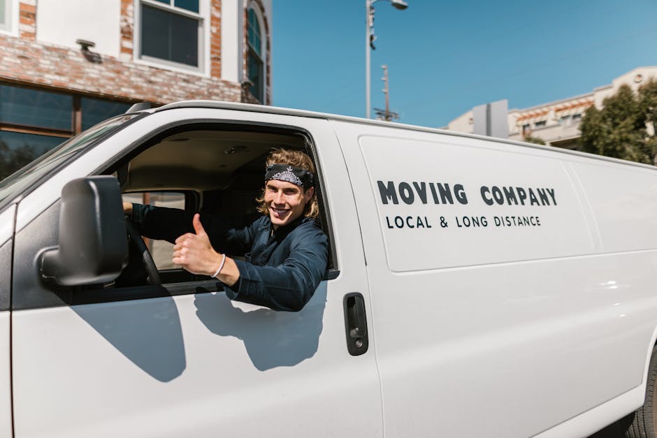 A smiling male mover with long hair tied back, wearing a dark blue shirt and a patterned headband, is sitting inside the cab of a white moving van with the text 'MOVING COMPANY LOCAL & LONG DISTANCE'
