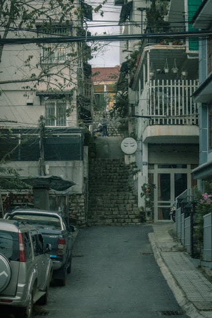 The image depicts a narrow urban alleyway leading up a flight of stone stairs, situated between residential buildings. The alley is paved with asphalt, with parked cars on the left side, including a silver vehicle closest to the camera. On the right, a building with a glass door and window features a small balcony decorated with potted plants and a metal railing. Overhead, a network of electrical and telecommunication wires crisscrosses above, with some supporting fixtures attached to the buildings. The background reveals a slight incline with additional residential structures, some trees, and a cloudy sky. This scene is representative of a typical city street where house removals or furniture transport might encounter tight access and stairs, making it relevant to relocation services provided by companies like Man with Van Harold Park, especially when managing bulky items in constrained environments, such as narrow stairs or no lift situations.