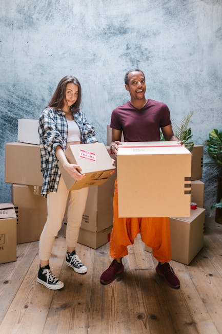 Two individuals, a woman and a man, are standing indoors in front of a textured grey wall, surrounded by stacked cardboard moving boxes of various sizes. The woman is wearing a checked shirt over a light top, beige trousers, and black-and-white sneakers, holding a medium-sized box with a red fragile sticker visible on it. The man is dressed in a maroon T-shirt, orange cargo shorts, and maroon shoes, carrying a larger cardboard box. The scene appears to depict a packing or furniture transport process, with the boxes arranged on the wooden floor, some partially opened or taped, indicating preparation for home relocation. The lighting is bright and natural, highlighting the clothing, boxes, and the overall setting associated with house removals and packing services, as provided by Man with Van Harold Park.
