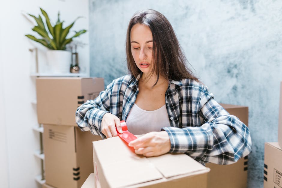 A young woman with long brown hair, wearing a blue and white checked shirt over a white top, is shown sealing a cardboard box with red packing tape. She is inside a room with a textured blue wall in the background, surrounded by several closed moving boxes of various sizes, some with printed labels. In the background, there is a white shelf holding a potted plant with long green leaves. The scene captures the packing process during a home relocation or furniture transport, with the woman preparing items for transportation. The environment suggests an organized approach to packing small flats on Romford High Street, with the attention to securing the boxes carefully before loading them onto a moving vehicle. The overall setting reflects the initial stages of a professional removals service, such as those provided by Man with Van Harold Park, associated with house removals and moving logistics.