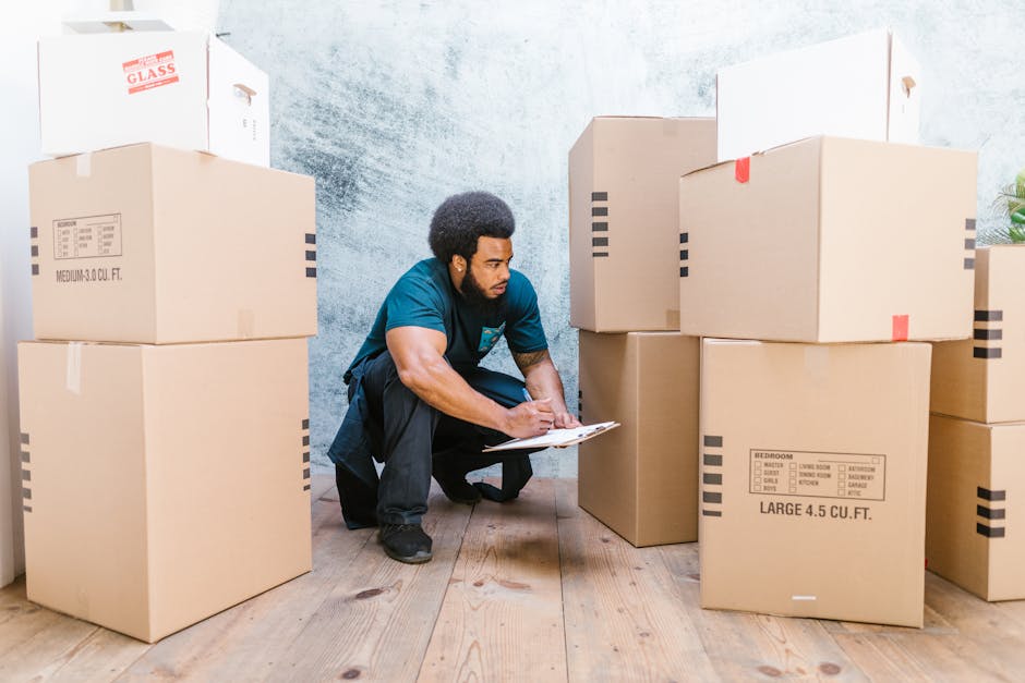 Two individuals, a woman and a man, are standing indoors in front of a textured grey wall, surrounded by stacked cardboard moving boxes of various sizes. The woman is wearing a checked shirt over a light top, beige trousers, and black-and-white sneakers, holding a medium-sized box with a red fragile sticker visible on it. The man is dressed in a maroon T-shirt, orange cargo shorts, and maroon shoes, carrying a larger cardboard box. The scene appears to depict a packing or furniture transport process, with the boxes arranged on the wooden floor, some partially opened or taped, indicating preparation for home relocation. The lighting is bright and natural, highlighting the clothing, boxes, and the overall setting associated with house removals and packing services, as provided by Man with Van Harold Park.