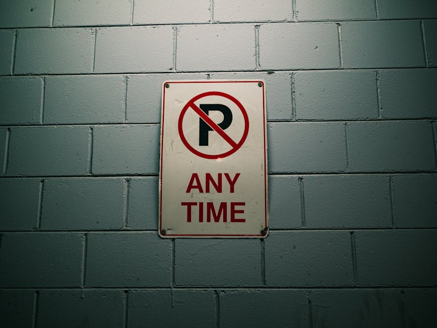 A close-up image of a rectangular metal sign attached to a black metal gate with vertical bars. The sign has a red background with white text that reads 'NO PARKING' at the top, followed by smaller te
